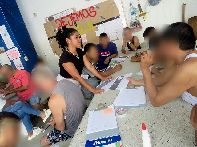 Hombres sentados alrededor de una mesa larga llenan formatos sobre tablas de apoyo mientras una de las practicantes les explica una hoja; al fondo, una cartelera hecha a mano. Las caras de los hombres están difuminadas.
