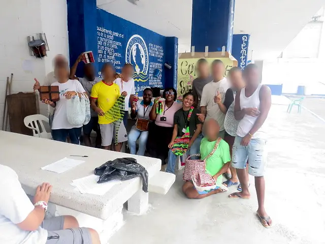 Un grupo de hombres y tres practicantes con carné posan sonrientes en un patio cubierto, frente a un mural azul. Varios muestran bolsos tejidos, una mochila y una caja de madera hechos a mano. Las caras de los hombres están difuminadas.